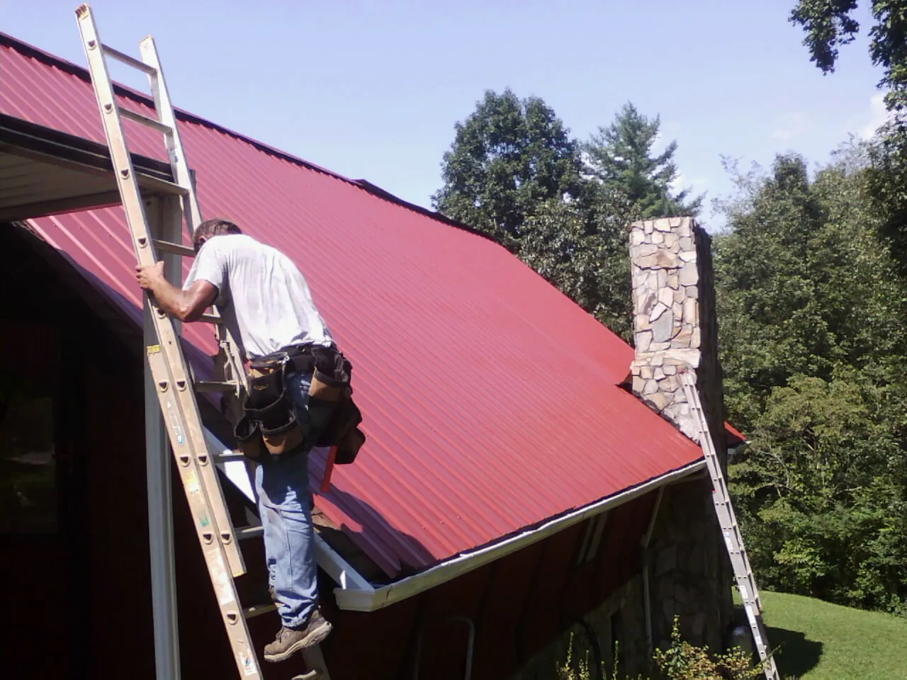 Roofer on a ladder beside a red metal roof and stone chimney — Soffit & Fascia Repair by Roofing Brothers in Kootenai